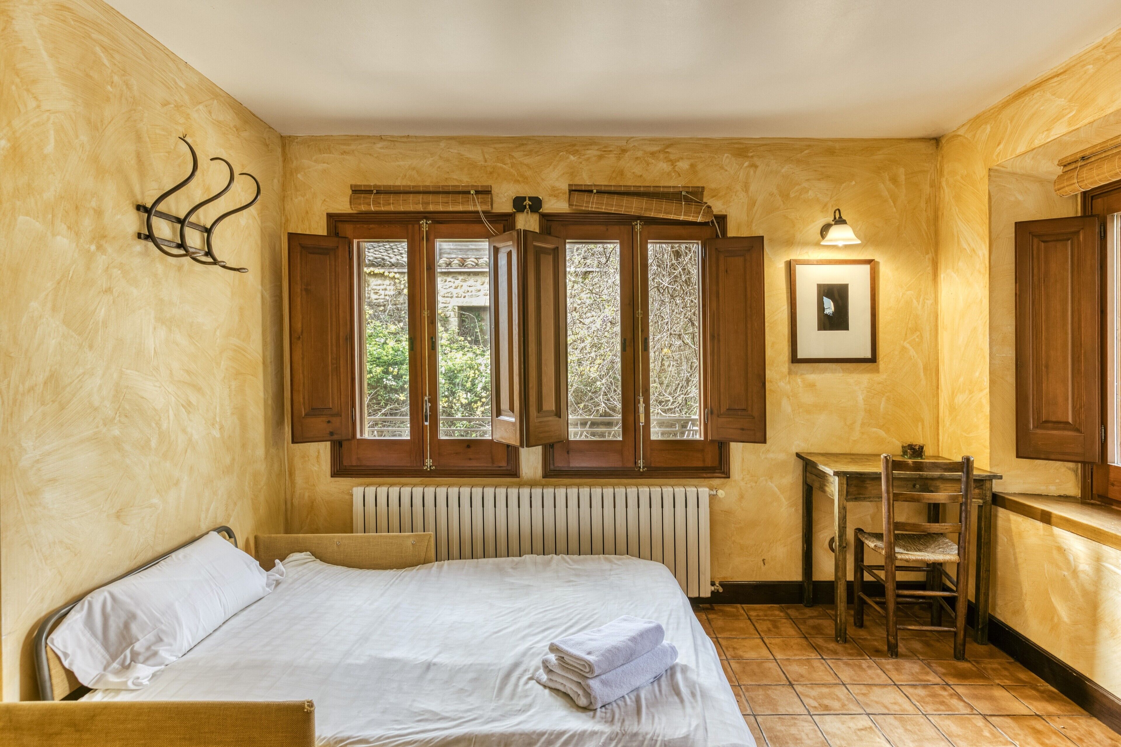 Une chambre à coucher comprenant un lit, une petite table, une chaise, un radiateur et une fenêtre avec des volets en bois.