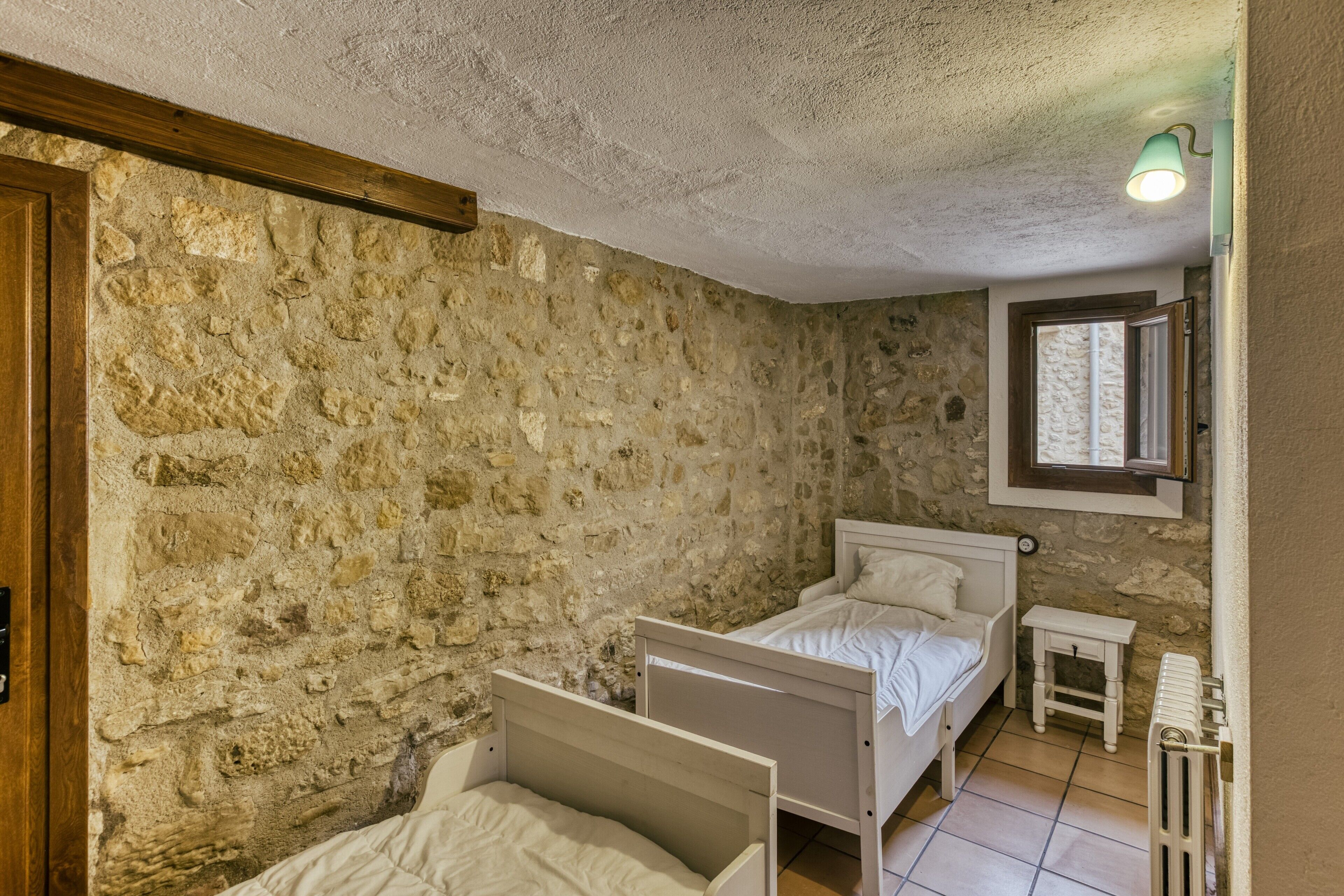 A room with stone walls, two beds, a small table, and a window with a view.