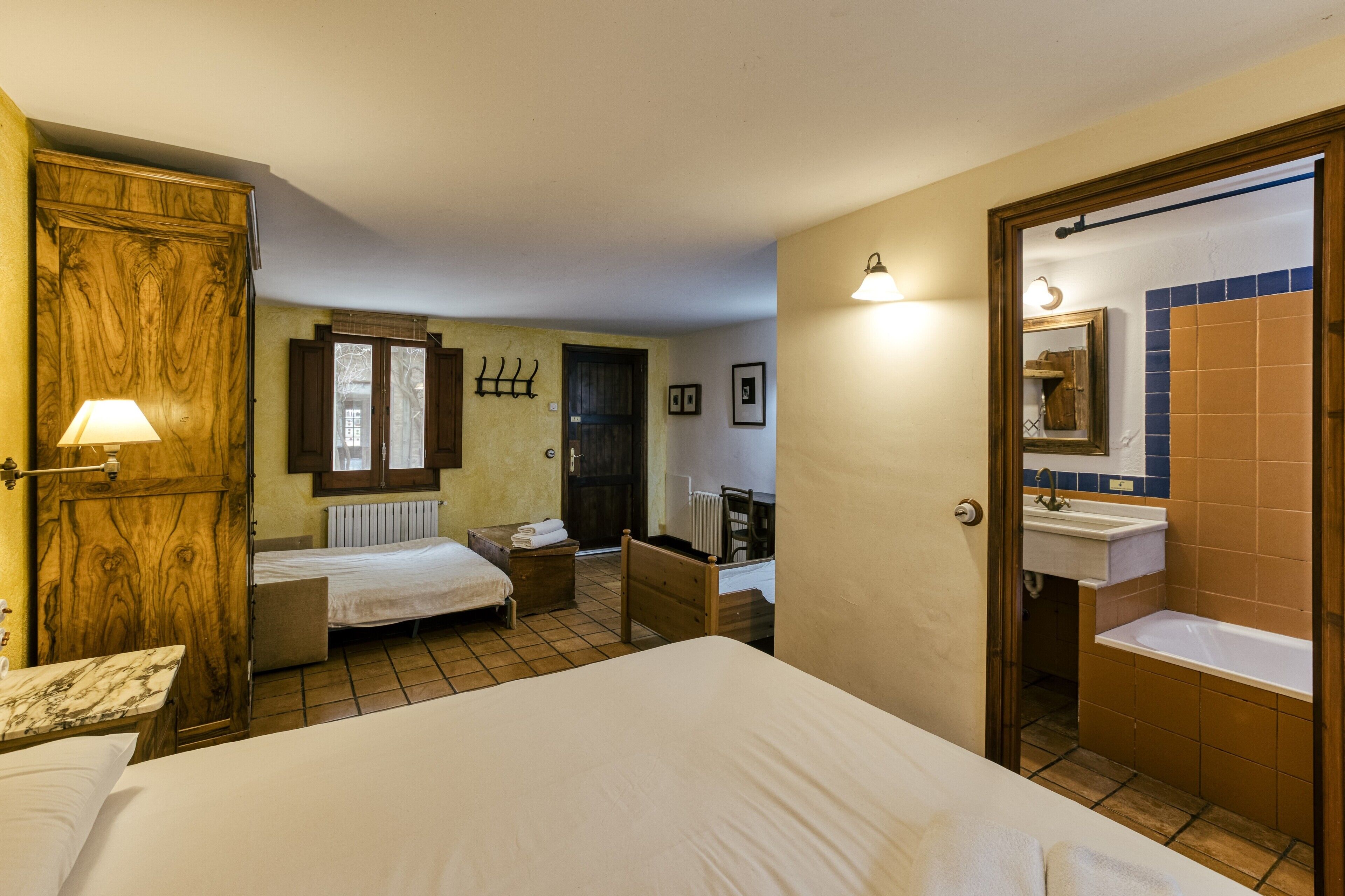A hotel room with a large bed, a wooden wardrobe, a small desk with a lamp, a bathroom with a sink and mirror, and a tiled floor.