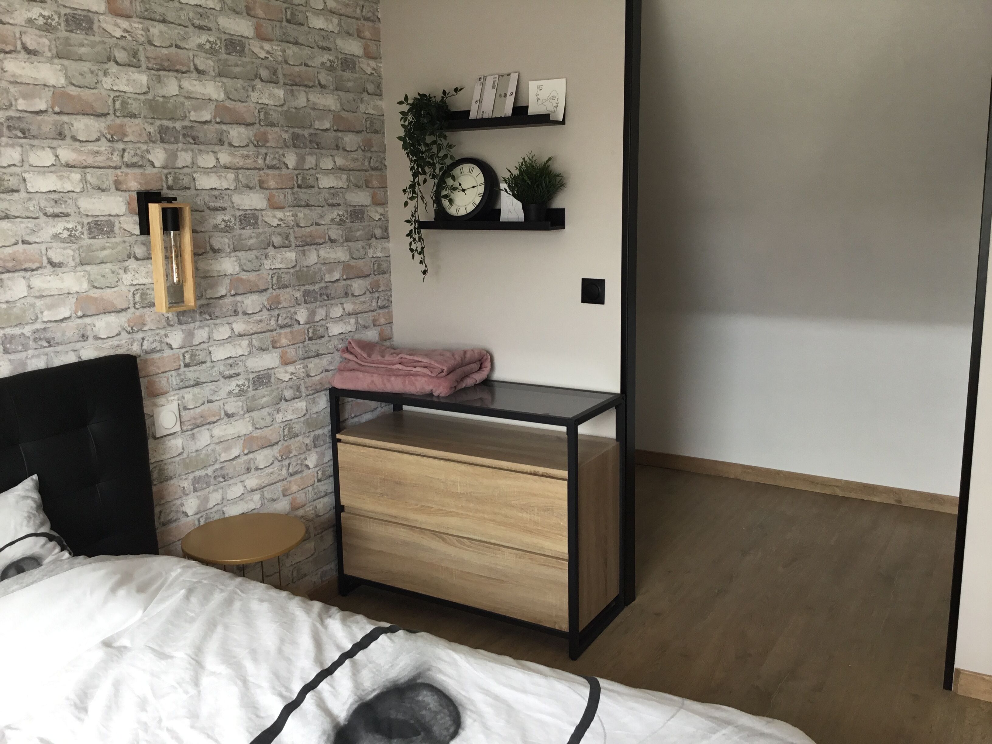 A bedroom with a brick wall, a bed with white bedding, a wooden nightstand with drawers, a small round table, and a shelf with a clock and plants.
