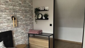 Un dormitorio con pared de ladrillo, una cama con ropa de cama blanca, una mesita de noche de madera con cajones, una pequeña mesa redonda y una estantería con un reloj y plantas.