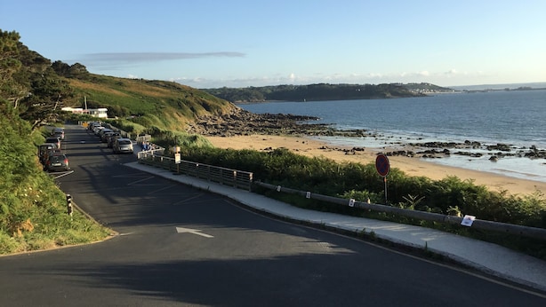 Una carretera costera con playa a la derecha, una zona de estacionamiento con coches, y cielo despejado.