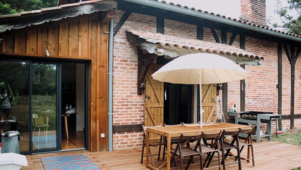A wooden deck with a table and chairs, a grill, and a brick building with large windows.
