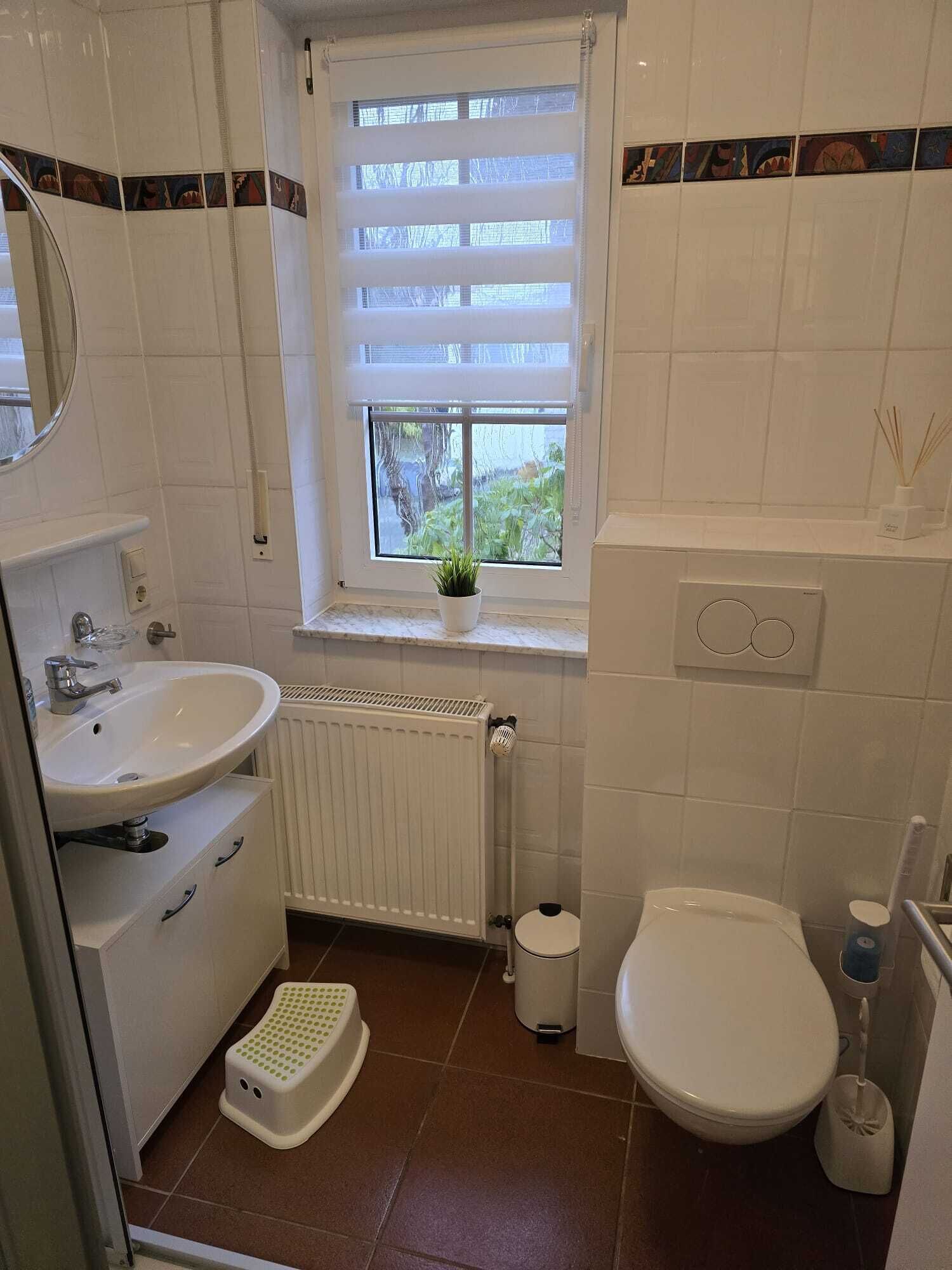 Ein Badezimmer mit weißem Waschbecken, Toilette und Heizkörper. Ein Fenster bietet Blick auf Grünflächen.