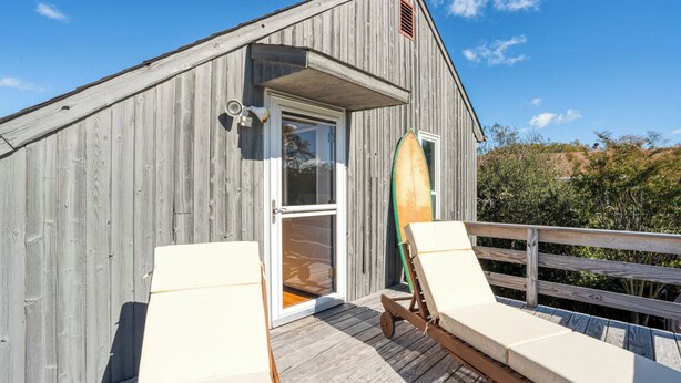 Un bâtiment en bois avec une porte, une planche de surf appuyée contre, et deux chaises longues sur une terrasse.