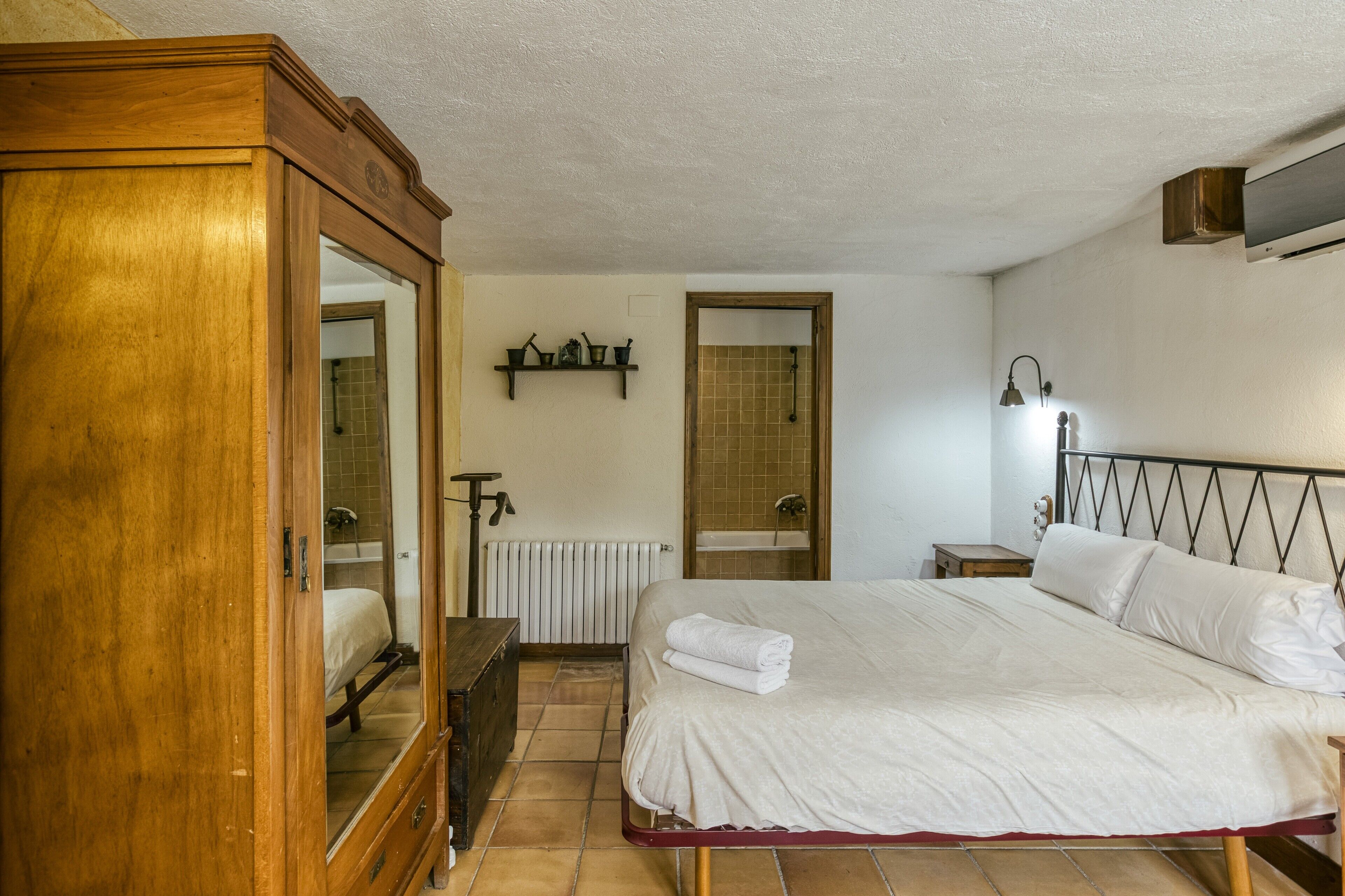 Un dormitorio con un armario de madera, una cama con ropa de cama blanca, un radiador, un estante con artículos de aseo y una lámpara de noche.