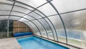 An indoor swimming pool with a curved glass ceiling, blue pool tiles, and a blue bench along the wall.