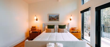 A modern bedroom with a large bed, two bedside tables, a bench with towels, and a view through a large window.