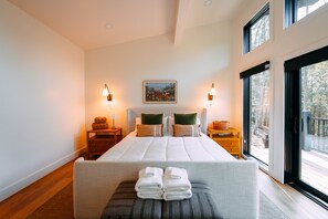 Un dormitorio moderno con una cama grande, dos mesitas de noche, un banco con toallas y vistas amplias por una ventana grande.