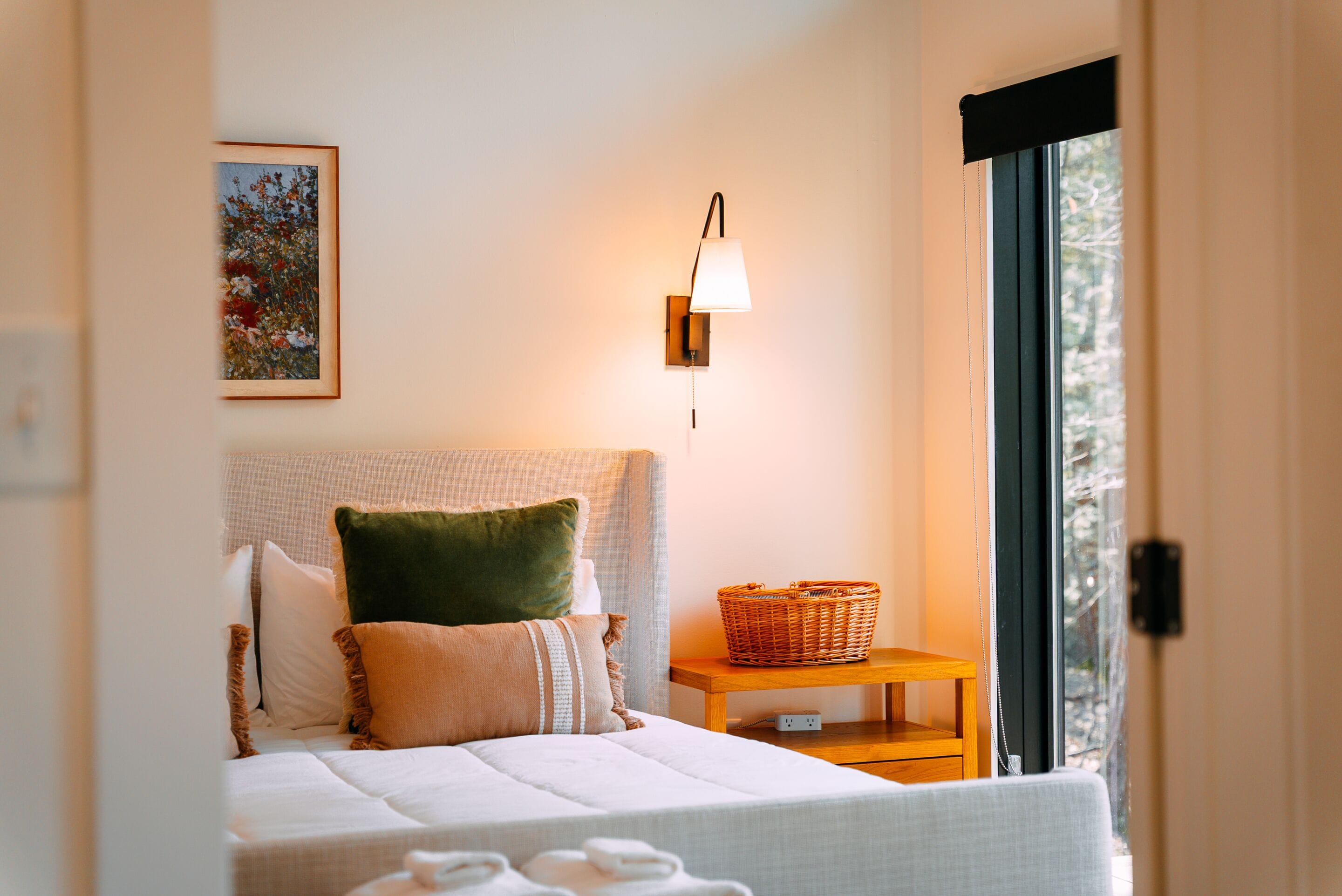 Une chambre à coucher bien rangée, avec un lit, des oreillers, une table de chevet sur laquelle se trouve un panier, une lampe fixée au mur et un tableau encadré.