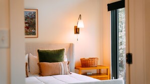 A neatly arranged bedroom with a bed, pillows, a bedside table with a basket, a wall-mounted lamp, and a framed picture.