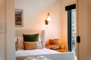 A neatly arranged bedroom with a bed, pillows, a bedside table with a basket, a wall-mounted lamp, and a framed picture.