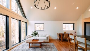 A spacious living area with a sofa, coffee table, dining table, and chairs, large windows, and a chandelier. - Maynard's Retreat | Hocking Hills, Modern, Hot Tub (Rockbridge)