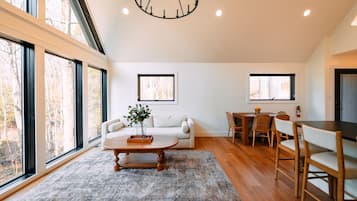 A spacious living area with a sofa, coffee table, dining table, and chairs, large windows, and a chandelier.