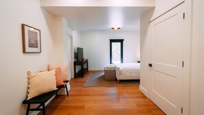 A room with a wooden floor, a white bed, a bench, a TV, and a window. - Maynard's Retreat | Hocking Hills, Modern, Hot Tub (Rockbridge)