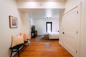 A room with a wooden floor, a white bed, a bench, a TV, and a window.