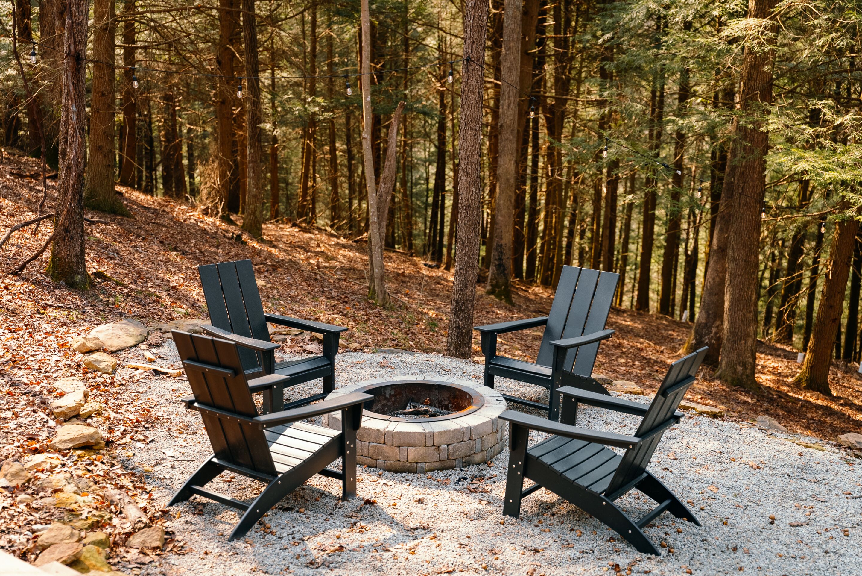 A circular fire pit surrounded by four Adirondack chairs in a forested area.