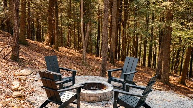 A circular fire pit surrounded by four Adirondack chairs in a forested area.
