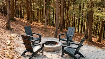 A circular fire pit surrounded by four Adirondack chairs in a forested area.