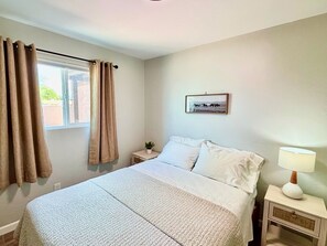 A bedroom with a bed, a nightstand, a lamp, a window with curtains, and a framed picture on the wall.