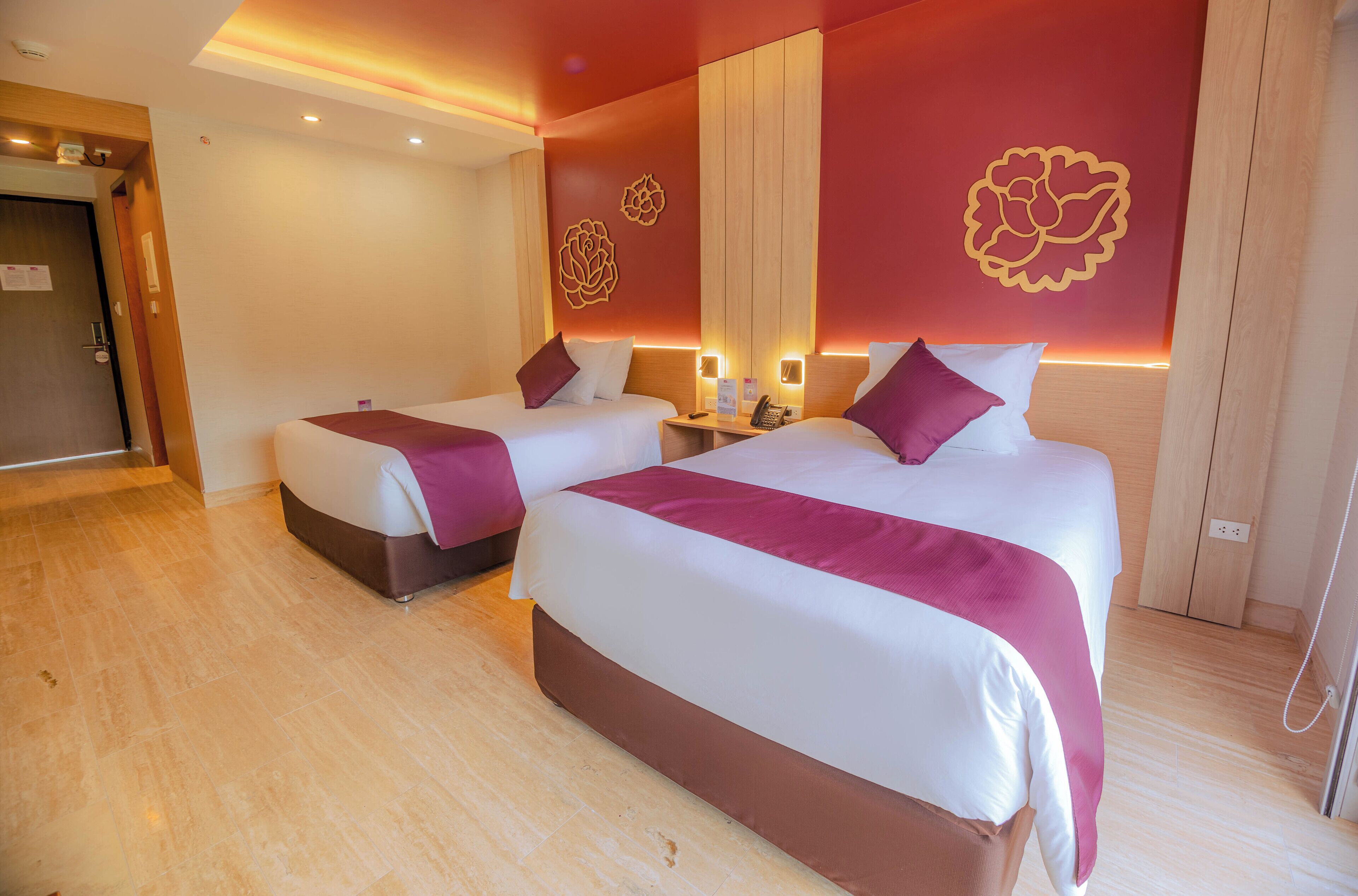 A hotel room with two beds, wooden flooring, and a red accent wall with floral designs.
