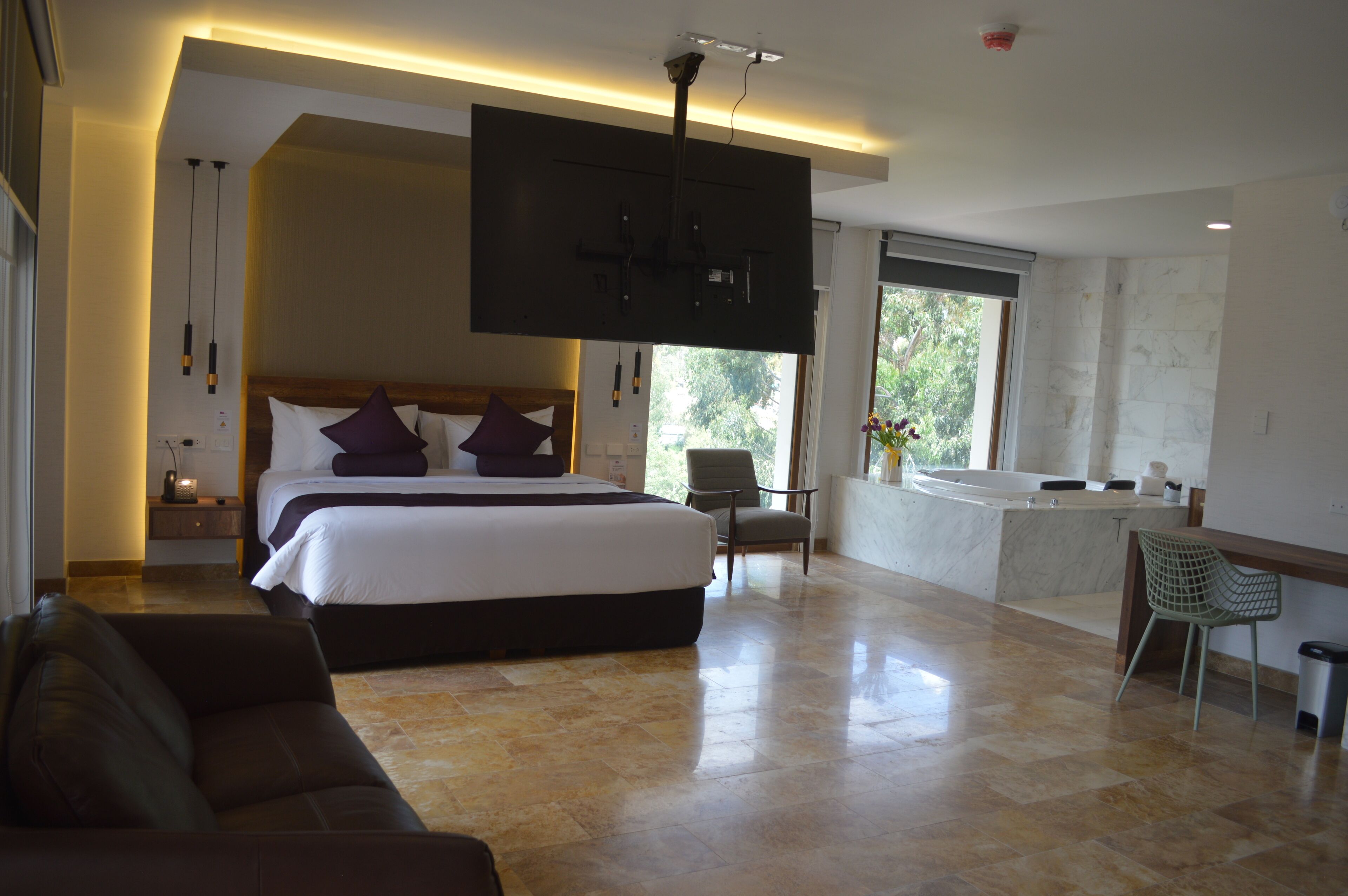 A spacious bedroom with a large bed, a sitting area with a sofa, a TV mounted on the wall, and a bathroom with a bathtub and sink.