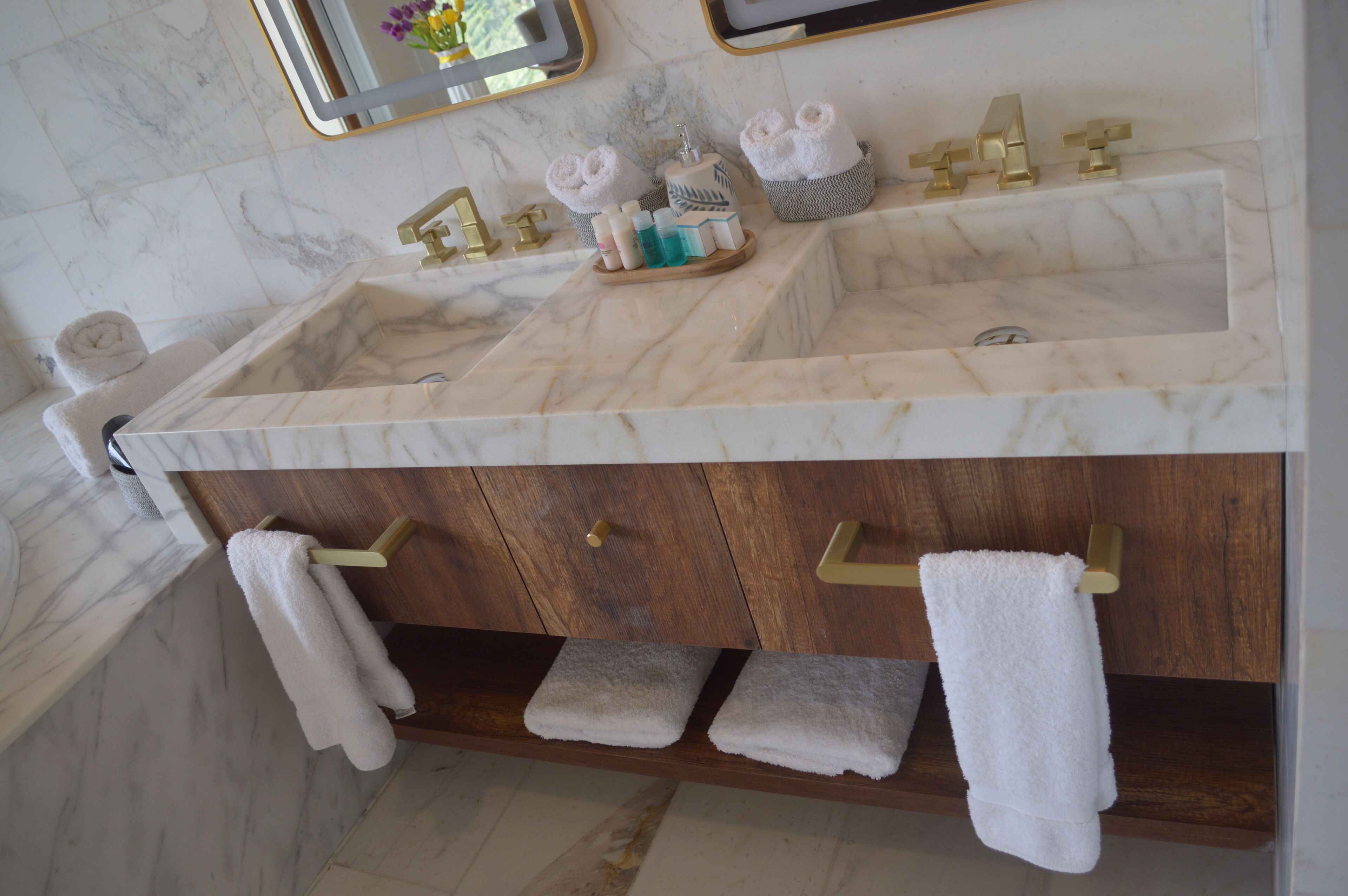 Une salle de bain avec deux vasques, des plans de toilette en marbre, des robinets dorés et des étagères en bois où sont posées des serviettes.