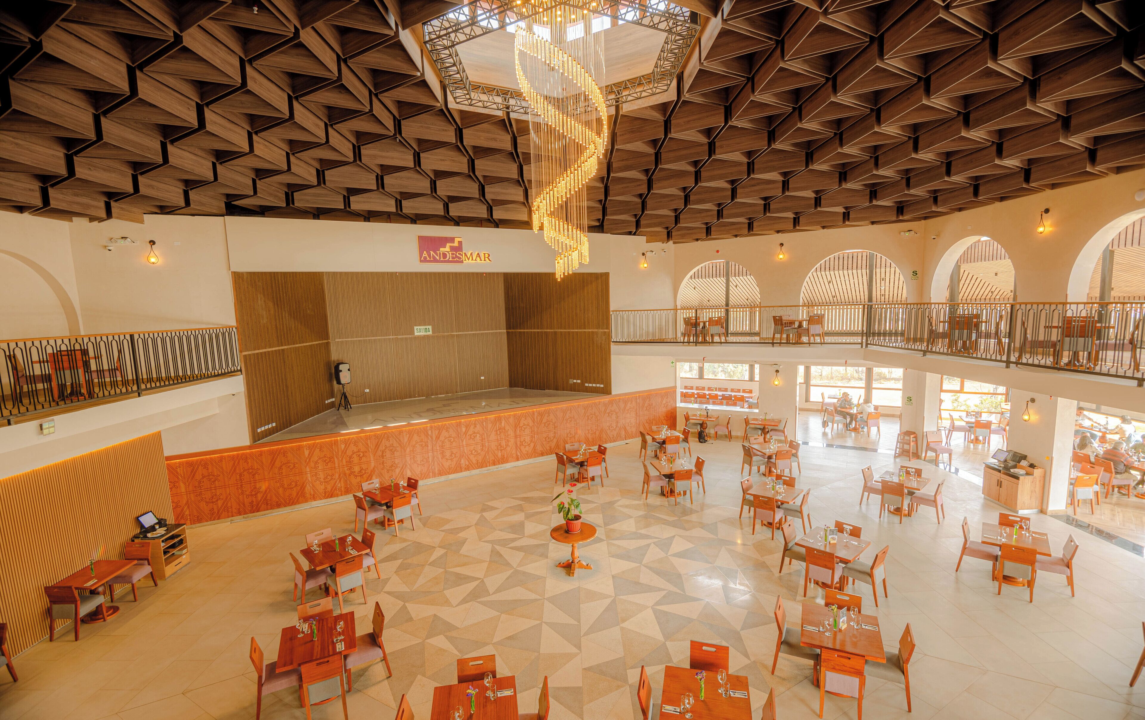 Un vestíbulo amplio con un escenario central, varias zonas de comedor y un techo alto con paneles de madera geométricos.
