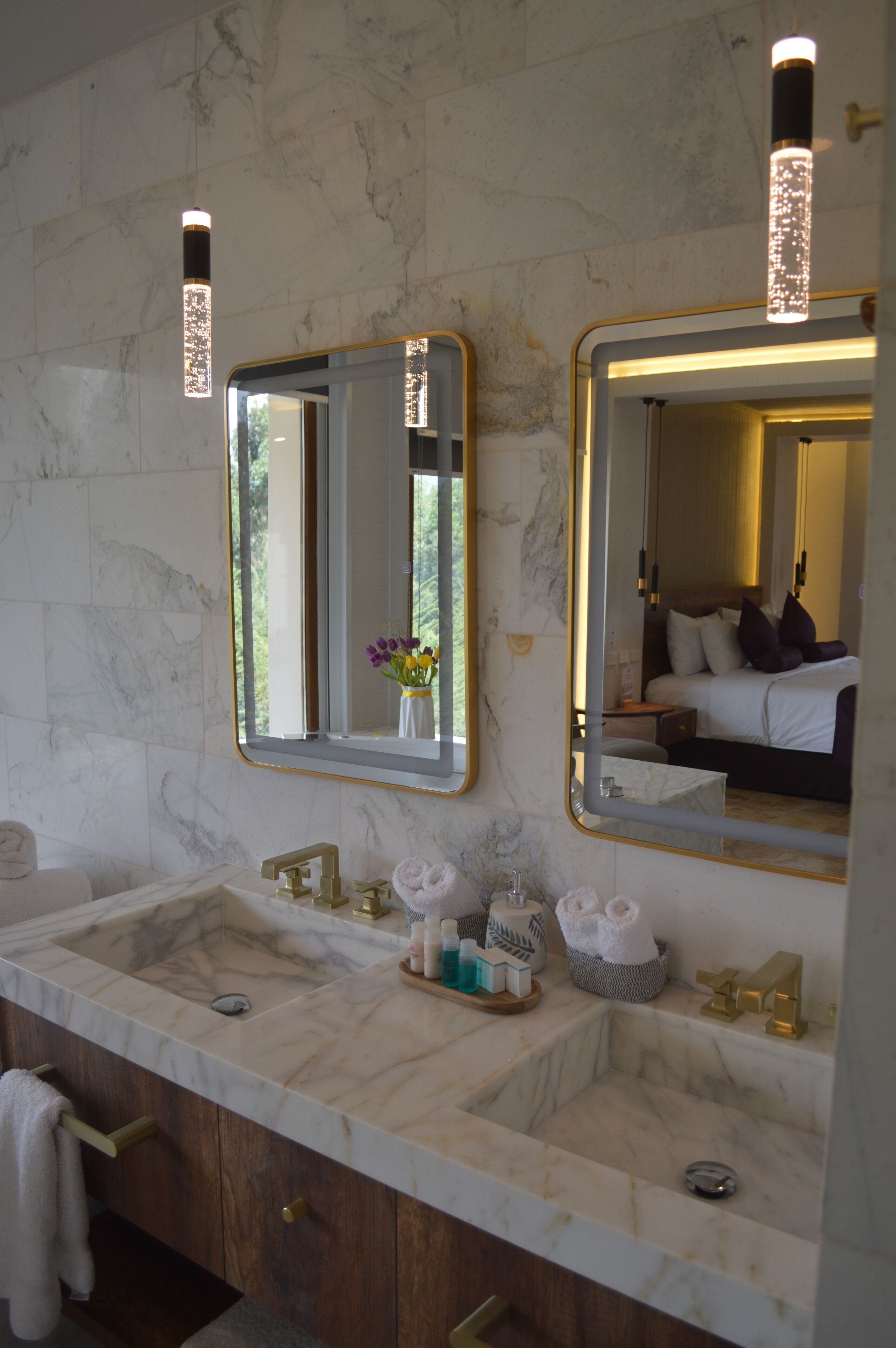 Un baño con dos lavamanos, paredes de mármol, accesorios dorados y un espejo que refleja un dormitorio.