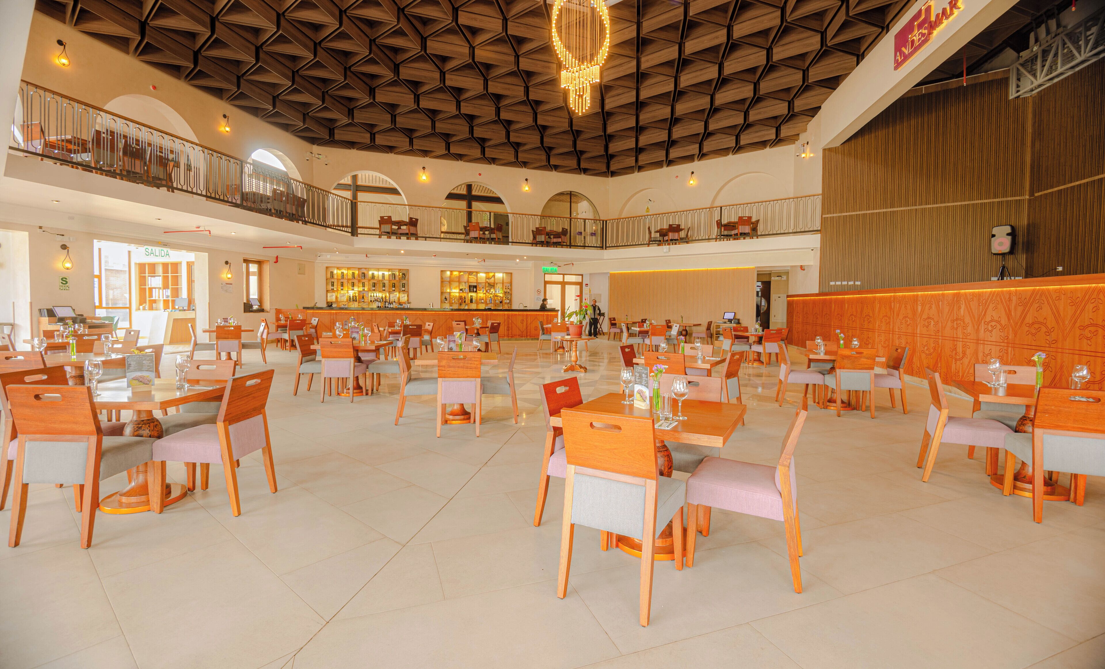 Een ruime hotel eetzaal met meerdere tafels en stoelen, een bar, en een hoog plafond met een geometrisch patroon.
