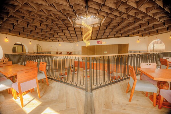 A spacious dining area with wooden tables and chairs, a central decorative light fixture, and a view of the dining area through a glass partition.