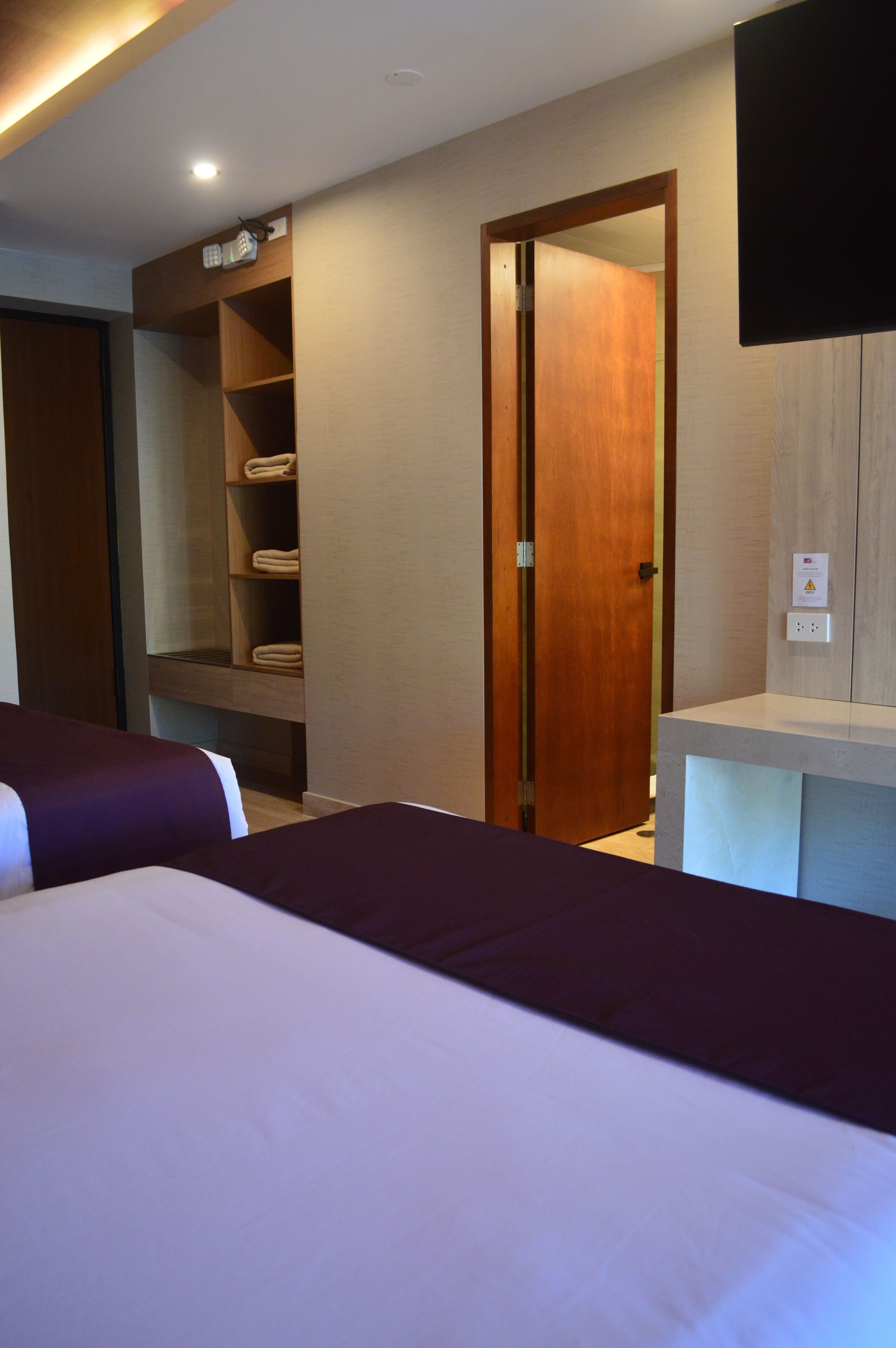 A hotel room with two beds, a wooden wardrobe, a shelf with towels, and a flat-screen TV mounted on the wall.