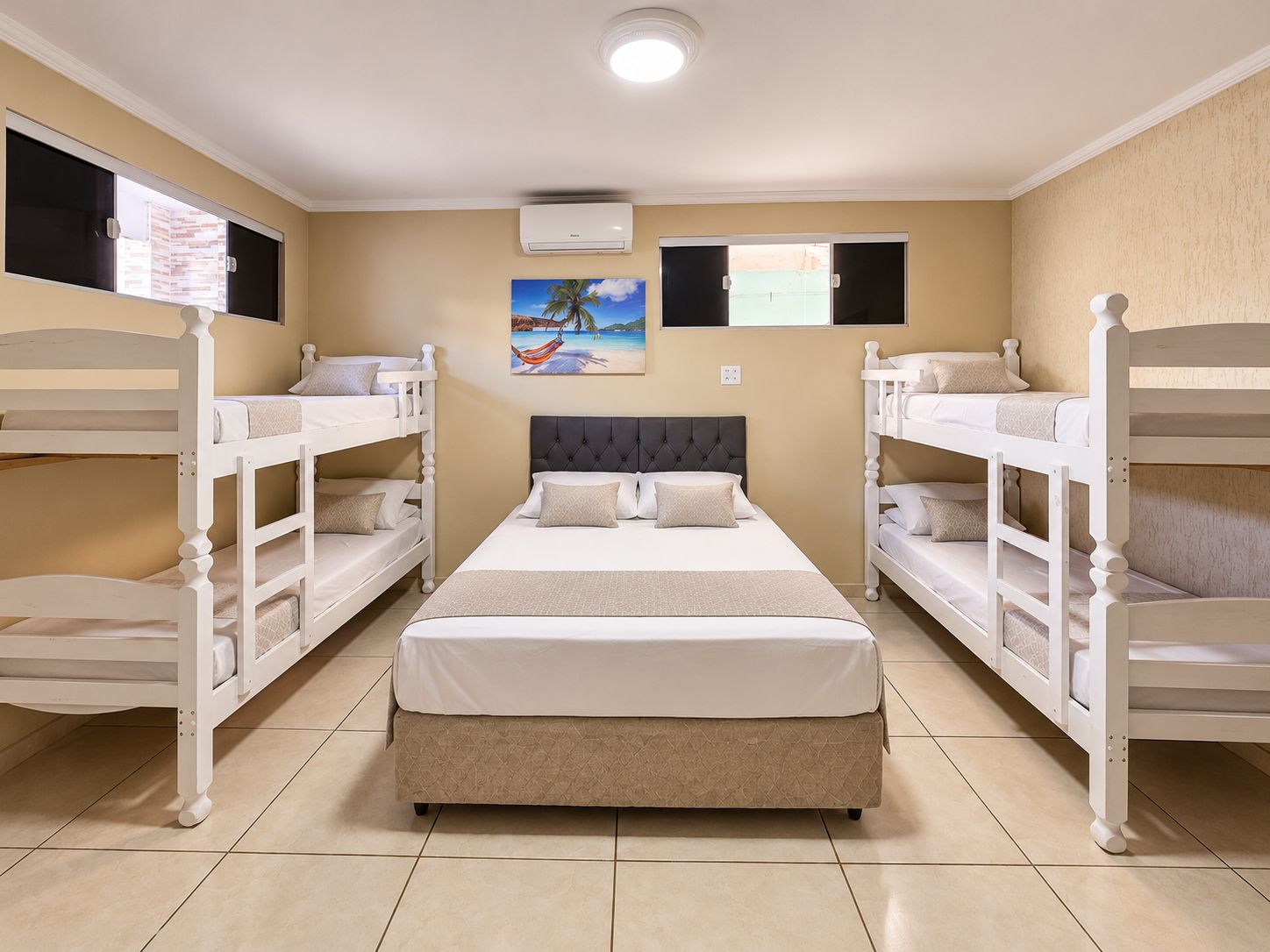 Une chambre d’hôtel avec un lit au centre, flanquée de deux lits superposés, un tableau au mur et un appareil de climatisation.