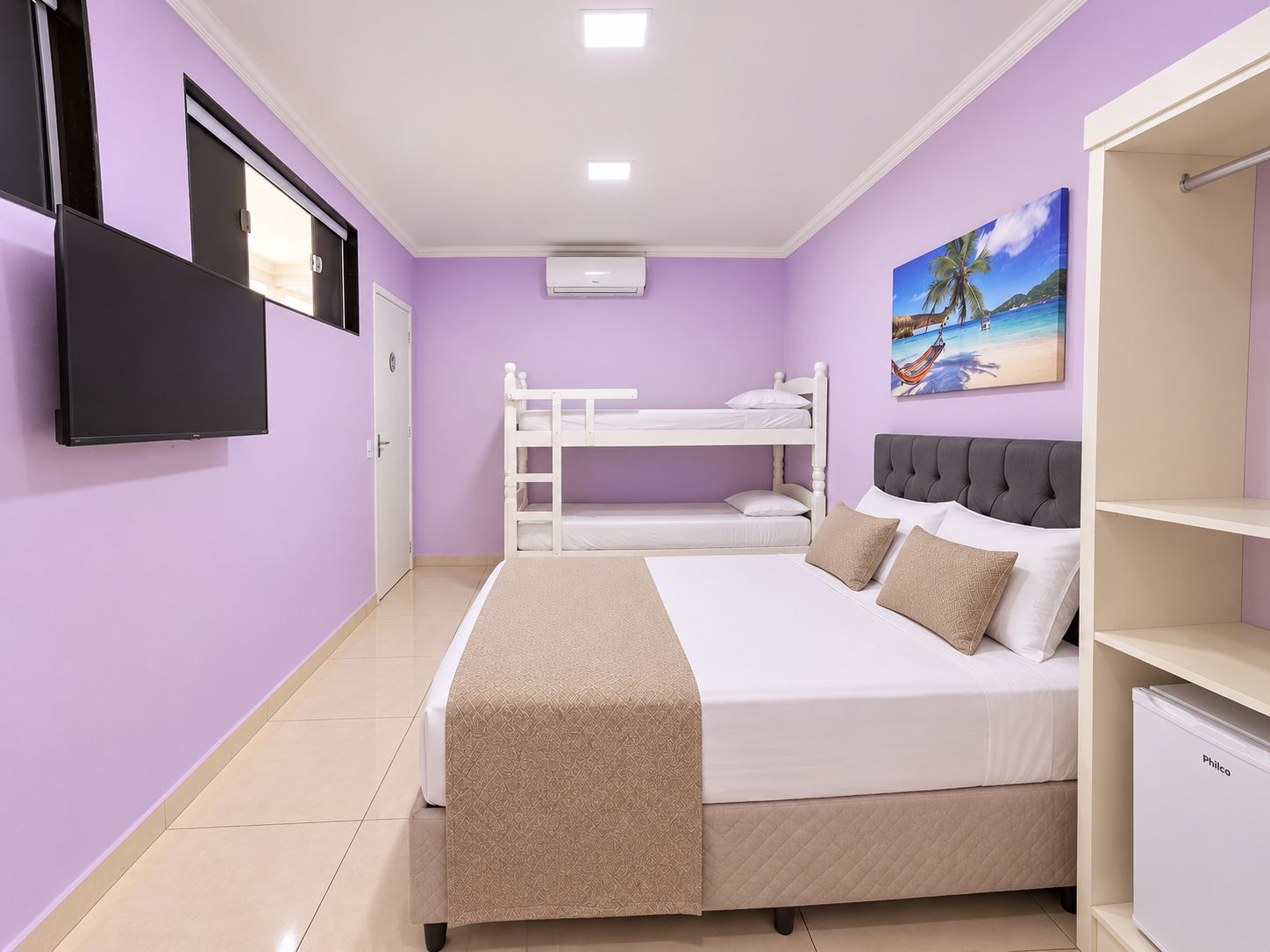 Une chambre avec un lit superposé, un lit simple avec une tête de lit, un téléviseur fixé au mur et un tableau représentant une scène de plage.