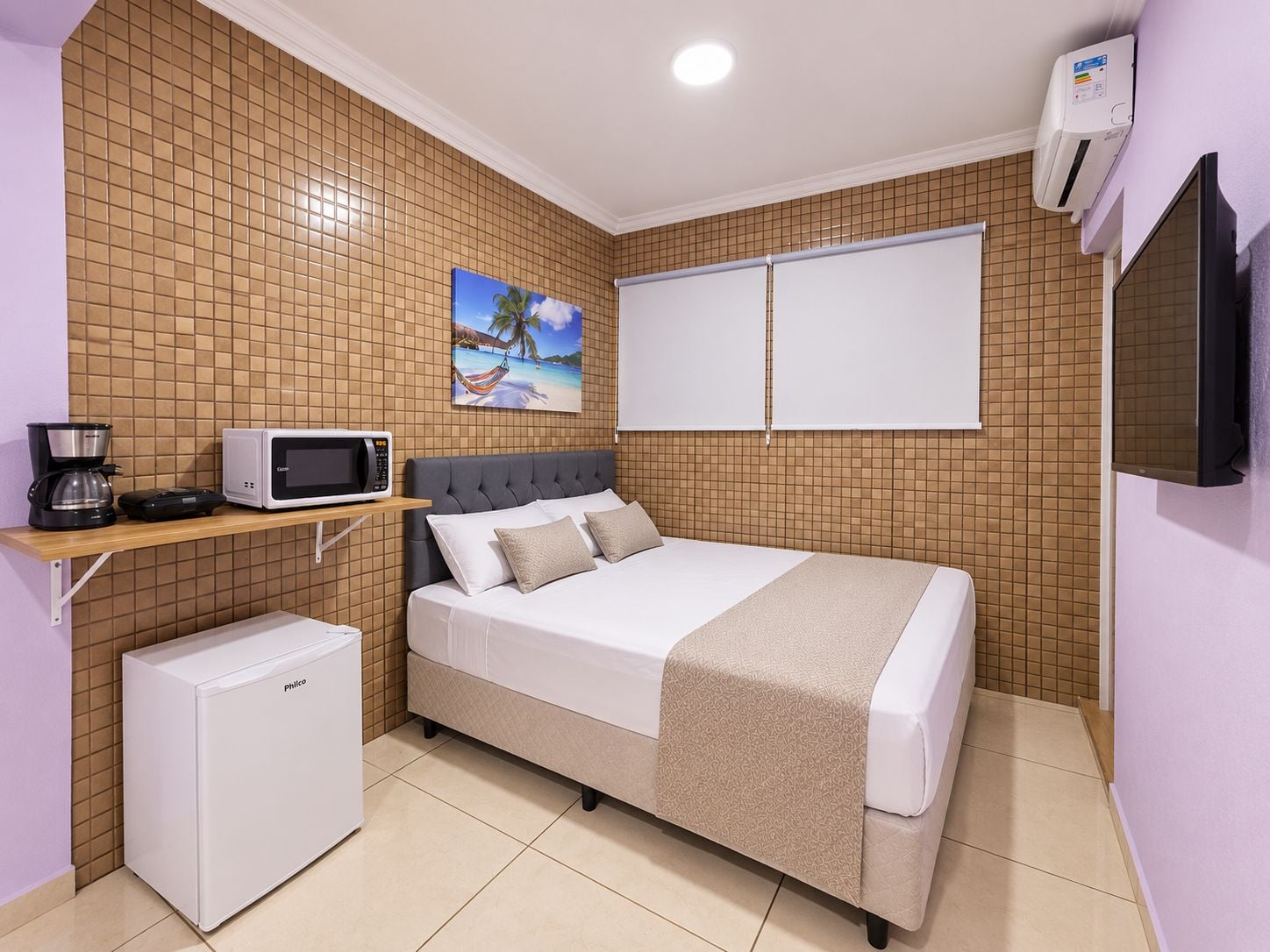 A compact hotel room with a bed, a small refrigerator, a microwave, a coffee maker, and a painting on the wall.