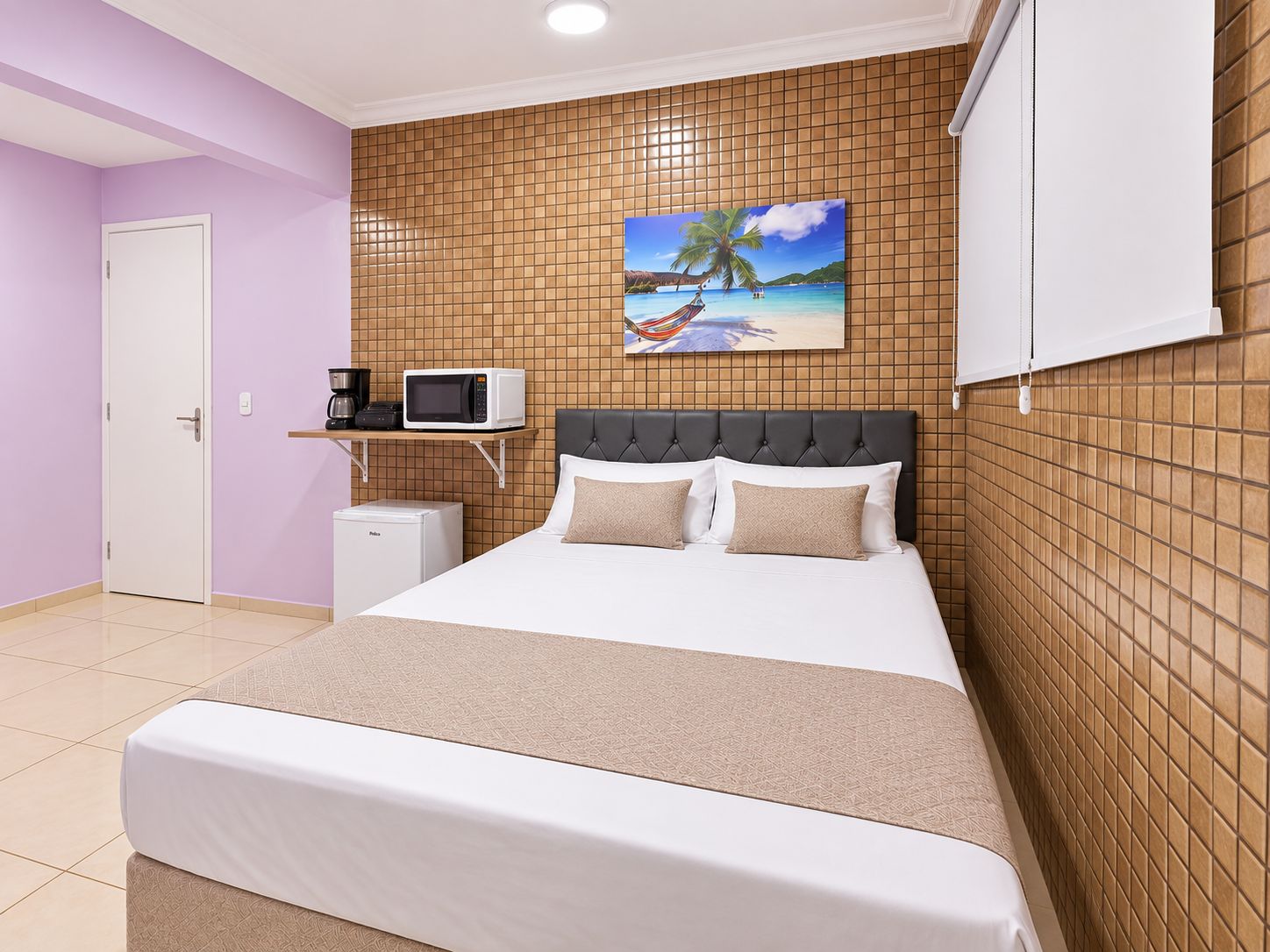 A hotel room with a bed, a microwave, a small fridge, and a painting of a beach scene.