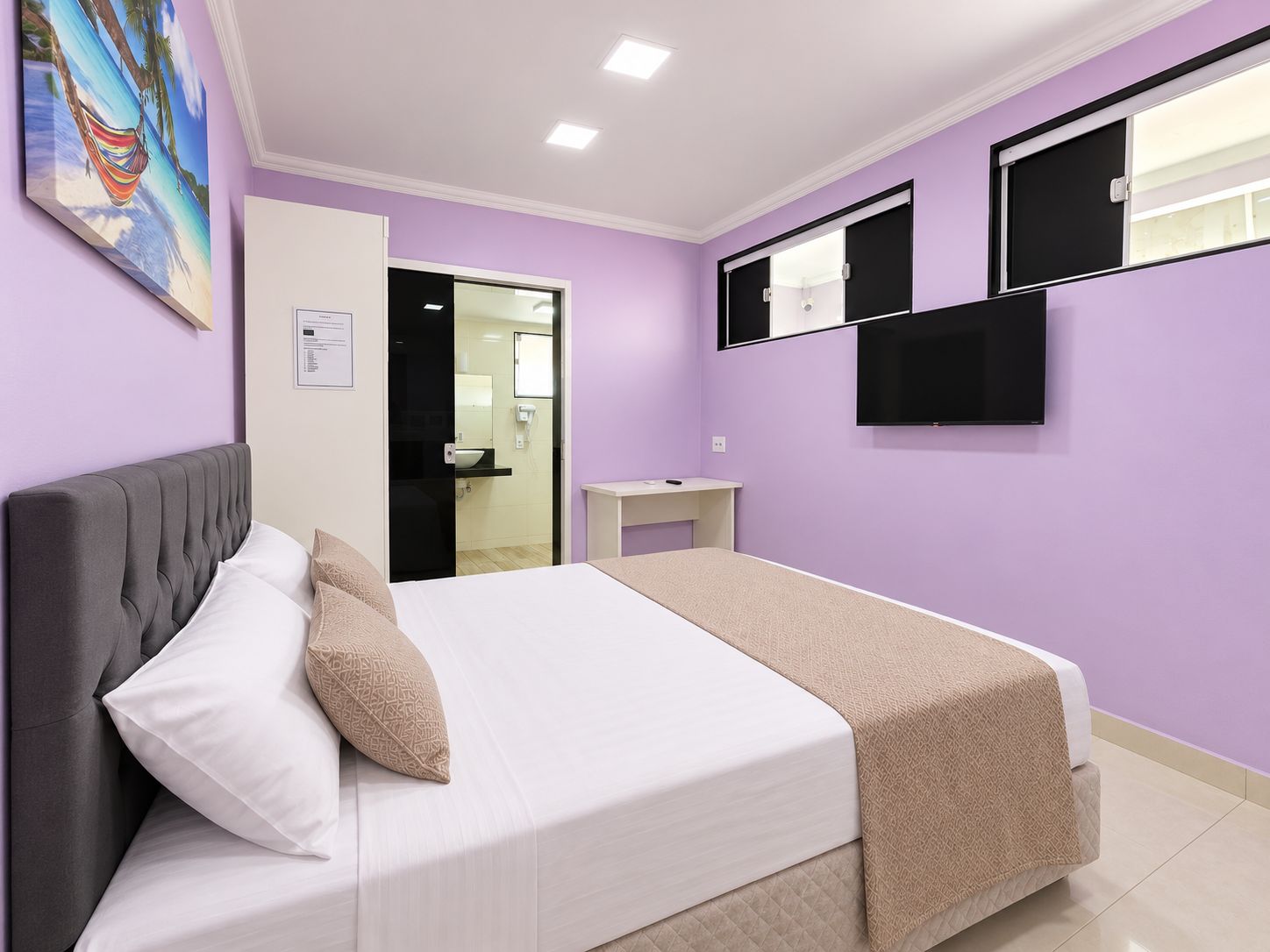 Une chambre à coucher avec un lit, une table de chevet, un téléviseur et une salle de bain visible à travers une porte ouverte.