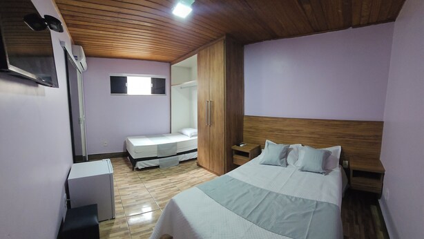 A bedroom with a wooden ceiling, a bed with white bedding, a nightstand, a wardrobe, and a small window. - Alice Suites (Arraial do Cabo)