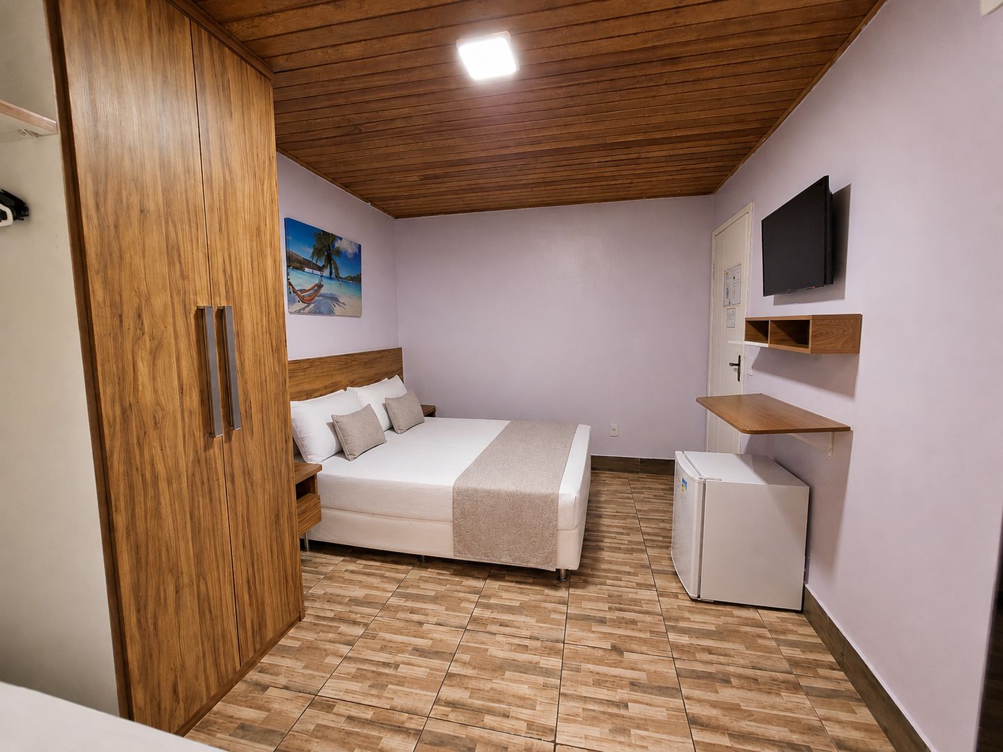 A bedroom with a bed, a wooden wardrobe, a television, and a small refrigerator.
