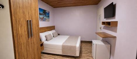 A bedroom with a bed, a wooden wardrobe, a television, and a small refrigerator.