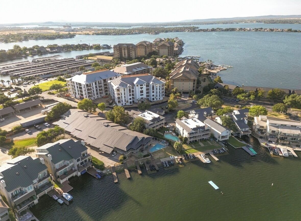 An aerial view of a waterfront property with multiple buildings, including a large white building with a swimming pool, surrounded by smaller structures and boats.