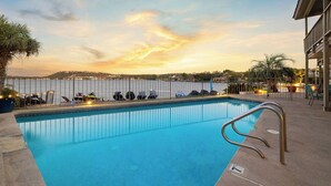 Una piscina con vistas al agua y una orilla lejana, rodeada de sillones de descanso y sombrillas.