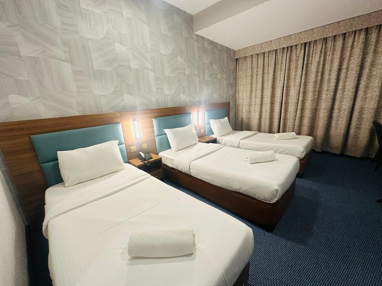 Habitació d'hotel amb tres llits, cadascun amb capçalera de fusta i roba de llit blanca. Hi ha taules de nit amb làmpades i un telèfon. La habitació té una paret amb un disseny i una catifa blava.