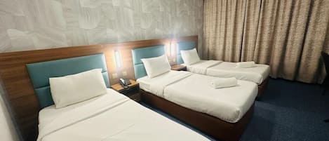 Habitació d'hotel amb tres llits, cadascun amb capçalera de fusta i roba de llit blanca. Hi ha taules de nit amb làmpades i un telèfon. La habitació té una paret amb un disseny i una catifa blava.
