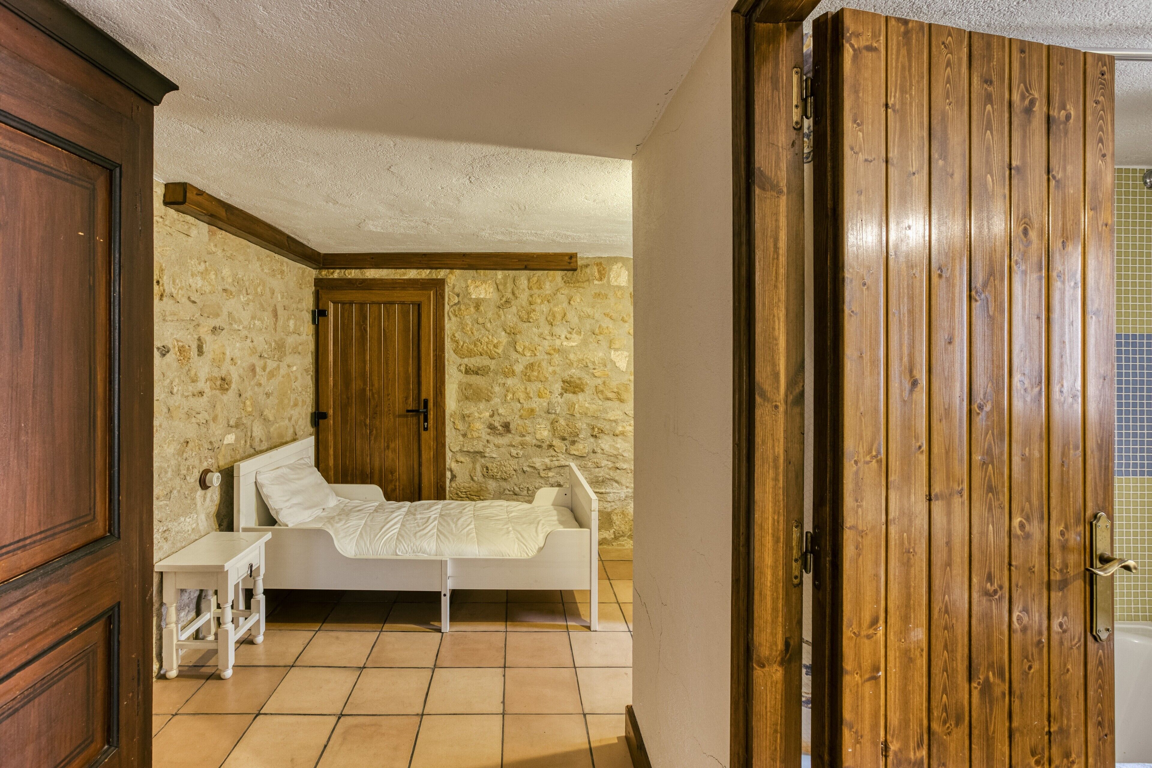 A bedroom with a stone wall, a wooden door, a white bed, and a small bedside table.