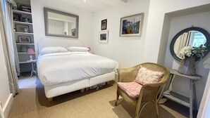 A bedroom with a bed, a wicker chair, a bedside table, a mirror, and a shelf with a vase. - Second nest Chelsea (London)
