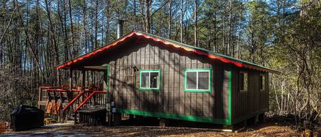 A wooden cabin with a red roof, green trim, and two windows with green frames, surrounded by a forest.