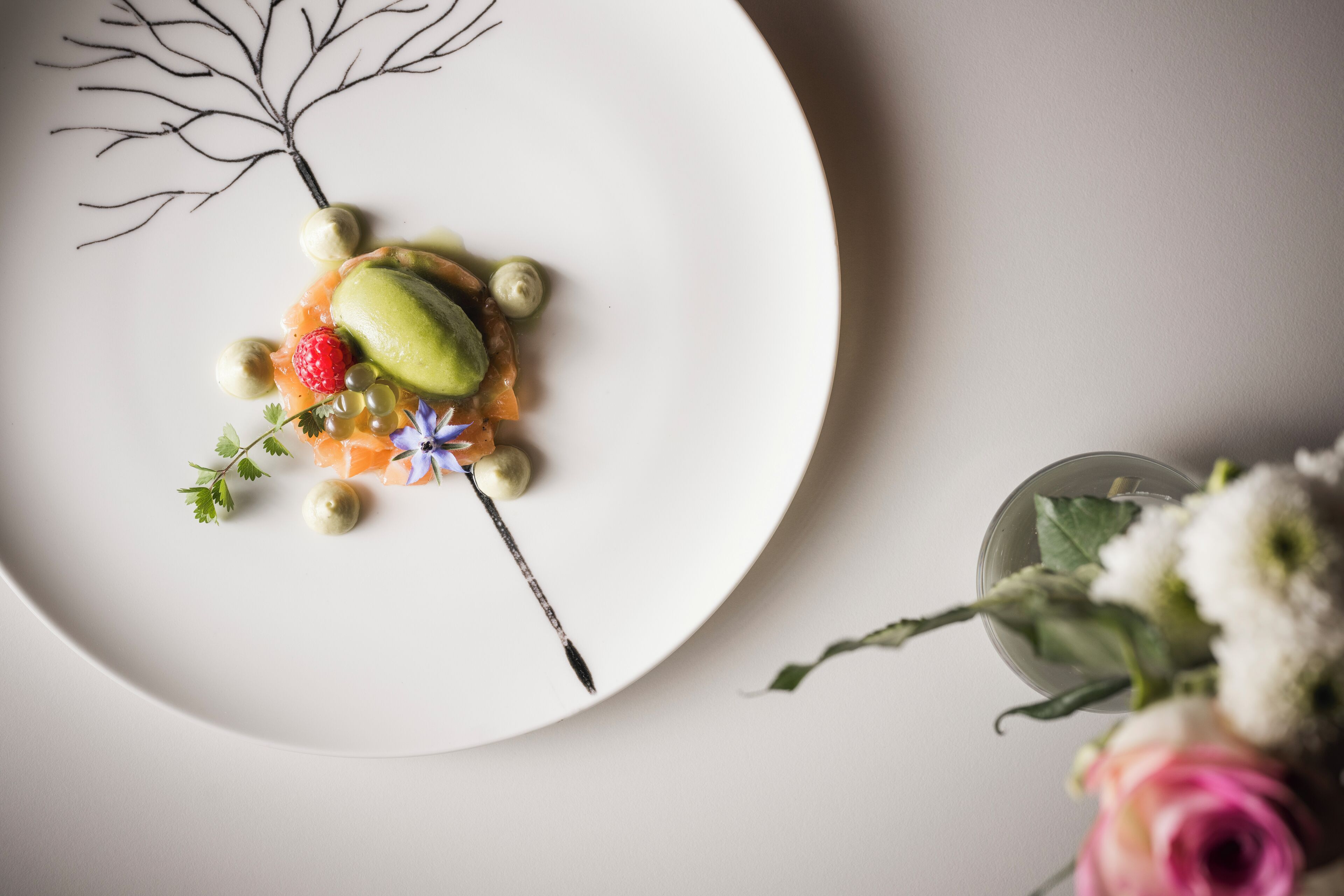Une assiette blanche sur laquelle est présentée une pâtisserie comprenant un ovale vert, des fraises et de petites boules, le tout accompagné d’une branche décorative et d’un petit verre d’eau dans lequel flottent des fleurs.