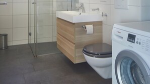 Moderni kylpyhuone, jossa on lasinen suihkuseinä, peilillä varustettu pesuallas, wc-istuin ja etukuormattava pesukone.