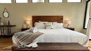 Een netjes ingerichte slaapkamer met een groot bed, houten hoofdbord, nachtkastjes met lampen, een klein tafeltje met fles en glazen, en een bankje.